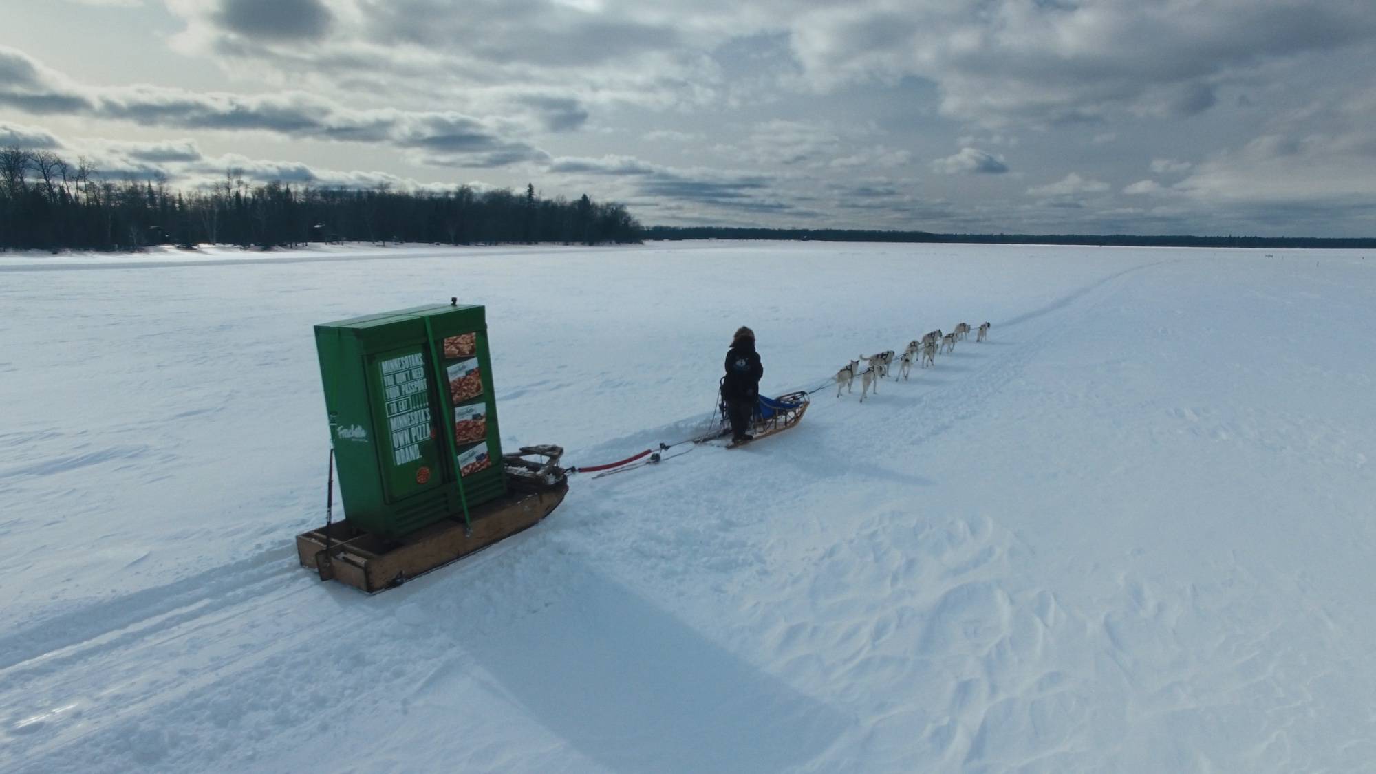 A dog sled pulls a giant freschetta pizza freezer over a frozen lake