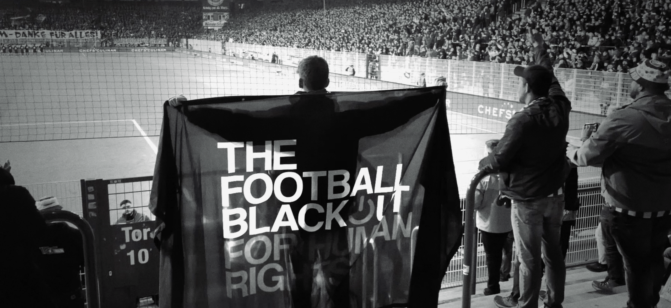 Man wearing flag at football game that reads "The Football Blackout For Human Rights"